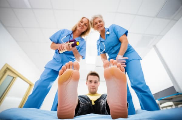 Create an image of two mature 40 year old women,  have their barefeet raised above a young looking 25 year old autistic  man with  short  brown hair and big blue eyes, have them crush/stomp him under their barefeet, a shrink ray gun is in the first woman's hands.  They are both grinning. Have the autistic man be looking up at their barefeet.

